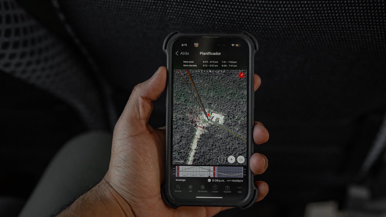 Mano che tiene uno smartphone con una mappa satellitare aperta su un’app di pianificazione, mostrando un punto segnato in un’area boschiva.